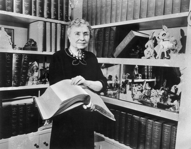 Helen Keller stands in a library holding an open book and smiling
