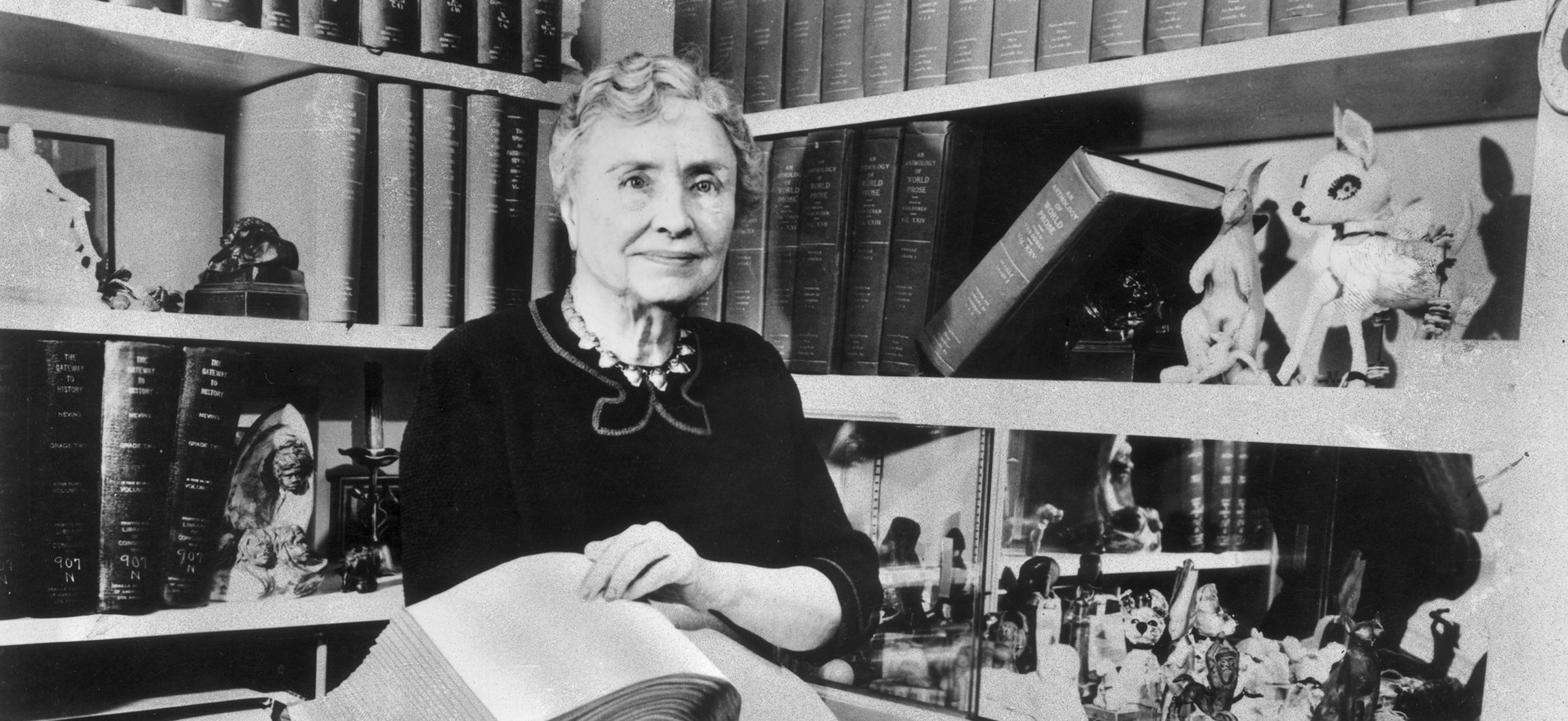 Portrait of American writer, educator and advocate for the disabled Helen Keller (1880 - 1968) holding a Braille volume and surrounded by shelves containing books and decorative figurines. A childhood illness left Keller blind, deaf and mute. (Photo by Hulton Archive/Getty Images)
