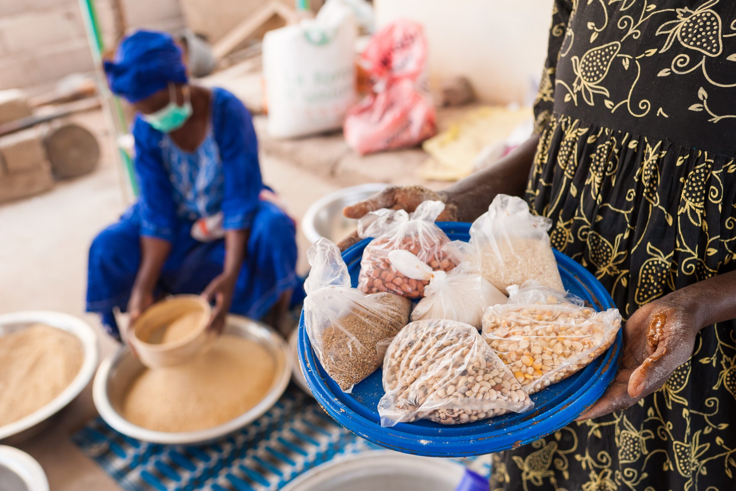 In the foreground, a woman holds a tray the ingredients used to make a nutrient-rich flour blend that is being sifted by the women in the background