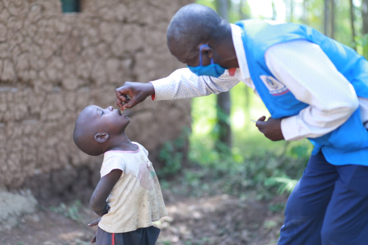 A health volunteer squeezes vitamin A drops into a young boy’s mouth while wearing a mask.