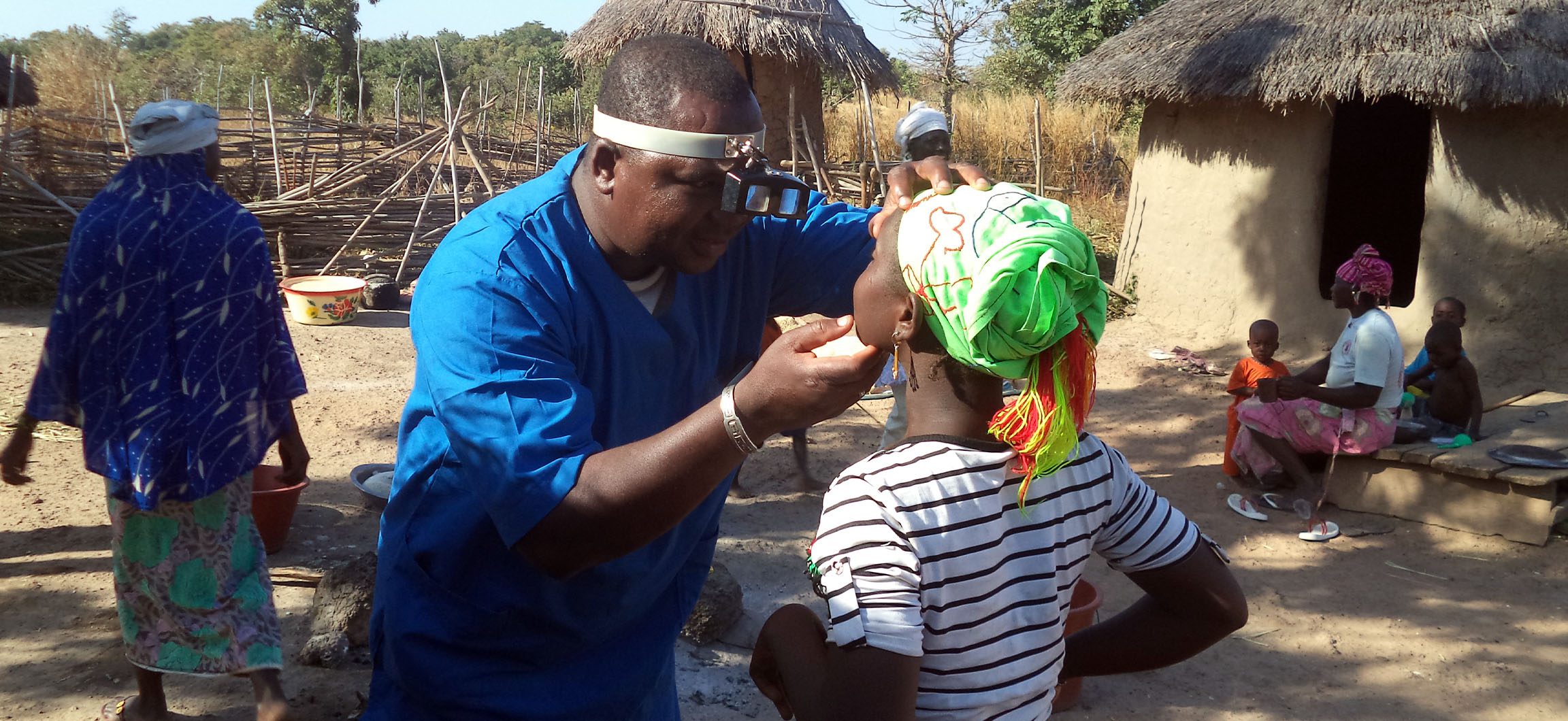 A healthcare worker examines the eyes of a young person. They are standing outside in a village.
