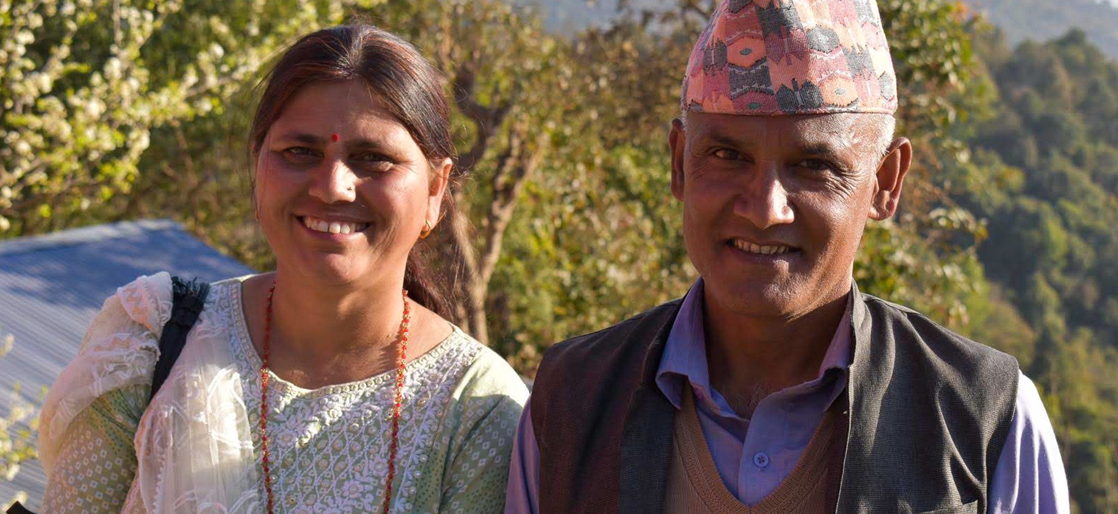 Gayatri and her husband, Bishwo, stand together smiling at the camera near their farm.