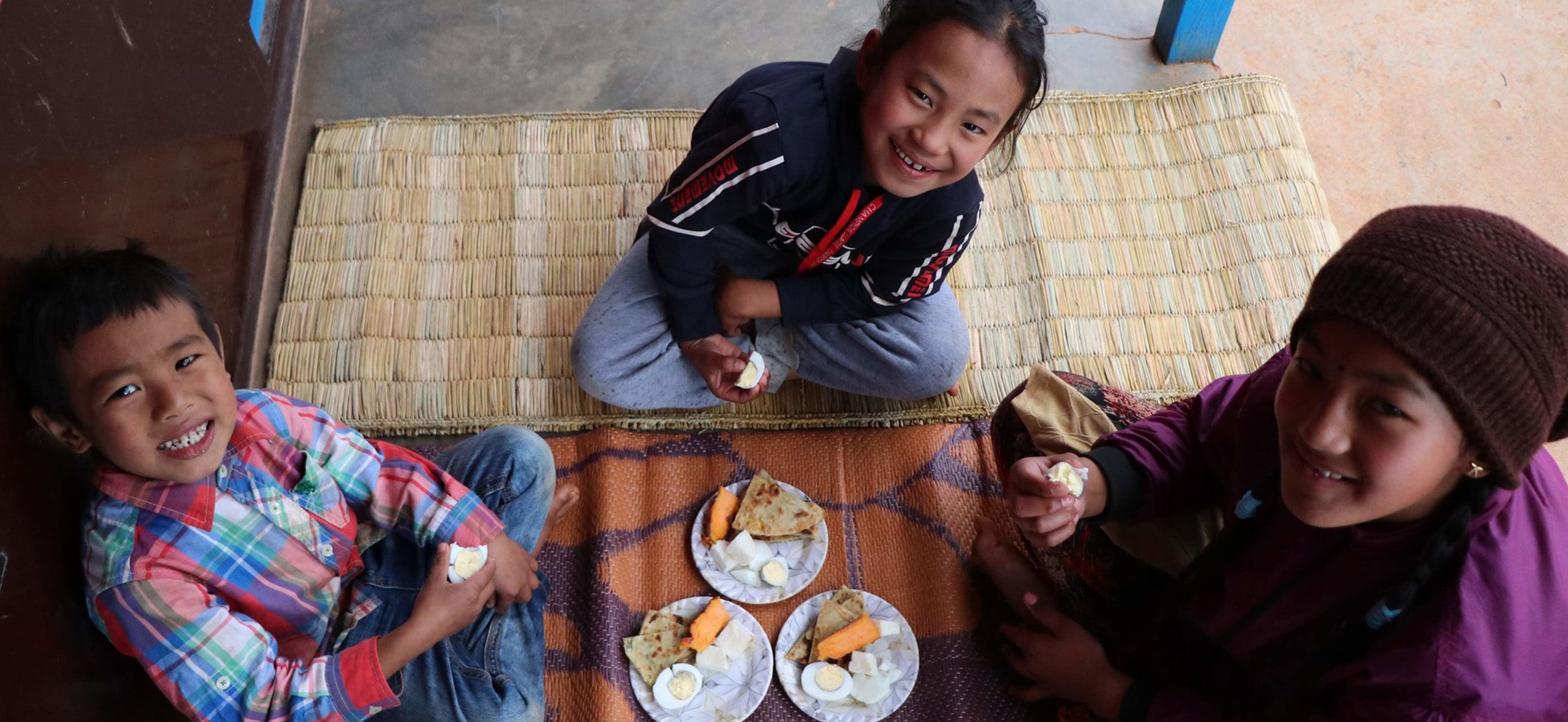 three smiling children sit on the floor together with nutritious food on the plates in front of them
