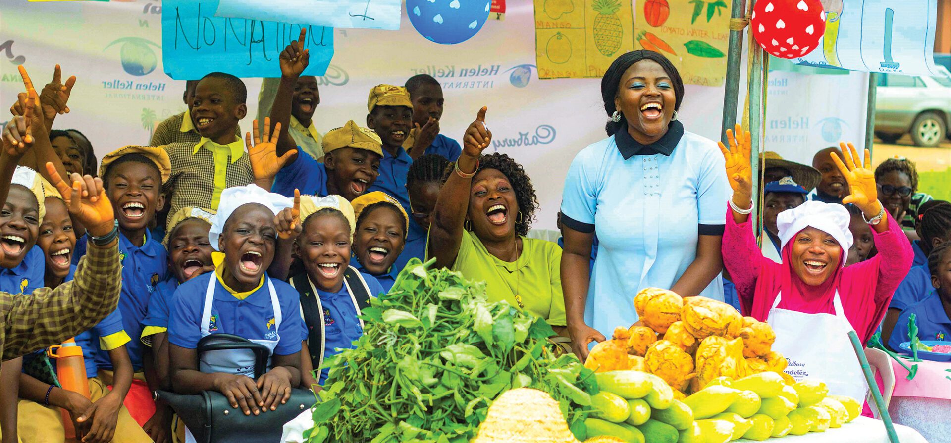 A classroom of female students and teachers cheer in front of a large pile of fresh vegetables.