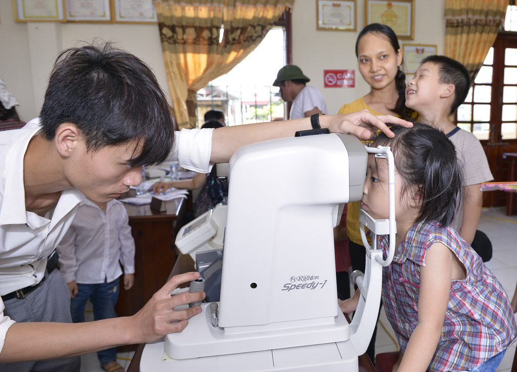 Optometrist uses autorefractor to check vision of young girl in clinic. Eye care professional performing eye exam with children waiting in background.