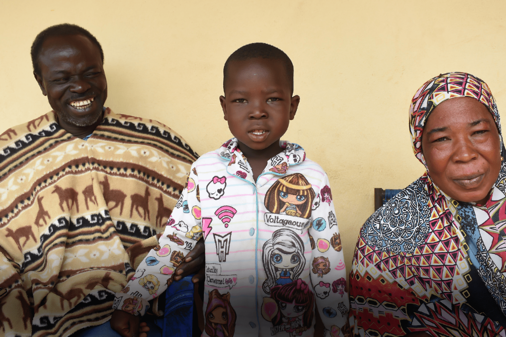 A photo of three people, Abdoul's father, Abdoul, and Abdoul's mother. Abdoul's father on the left is smiling towards Abdoul. Abdoul is looking at the camera with an open mouth and standing. Abdoul's mother is smiling closed-mouth to the camera.
