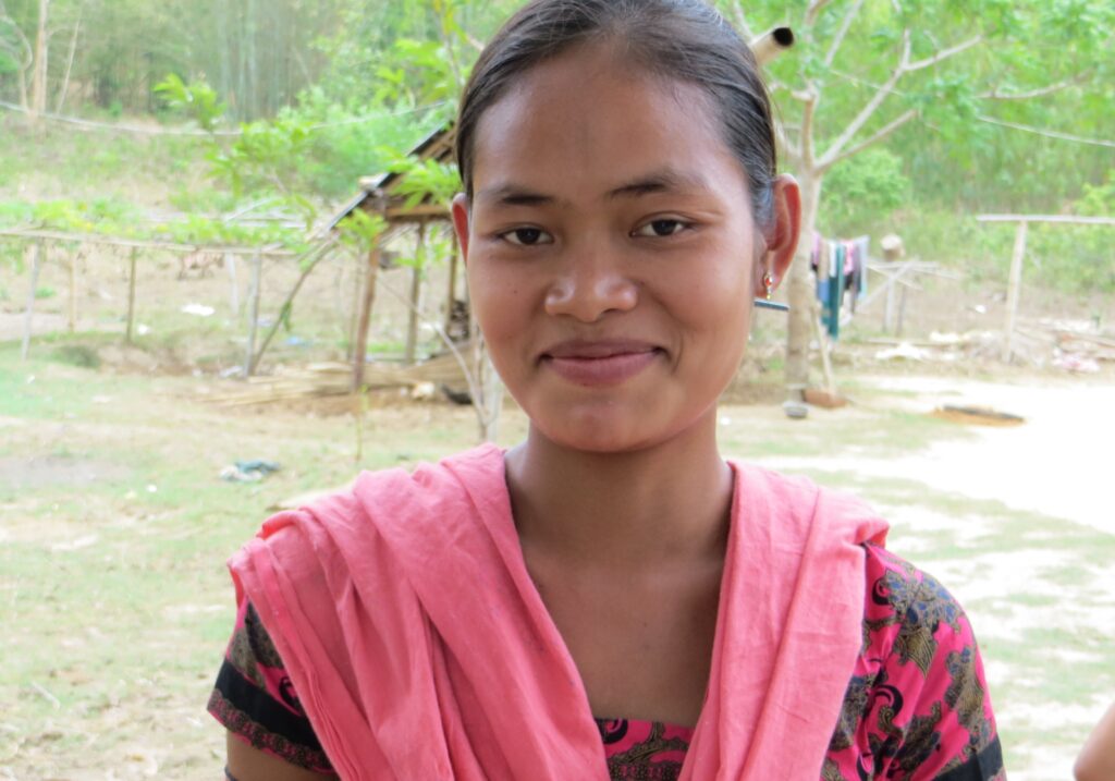 Smiling young woman wearing pink scarf and floral dress standing outside near a fence in a rural area.