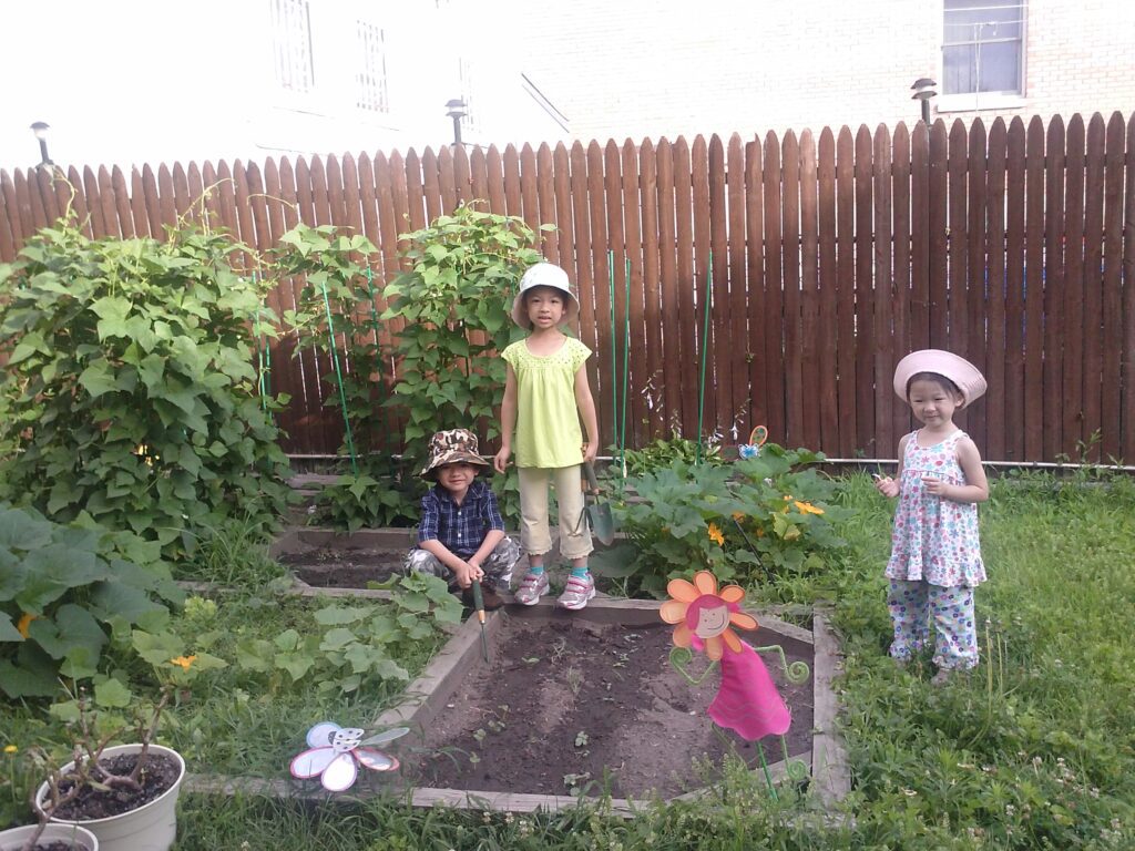 Three young children in hats are in a backyard garden with raised beds and green plants.