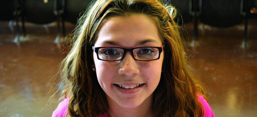 Close-up of a smiling young girl wearing glasses indoors. She has light brown hair and looks directly at the camera.