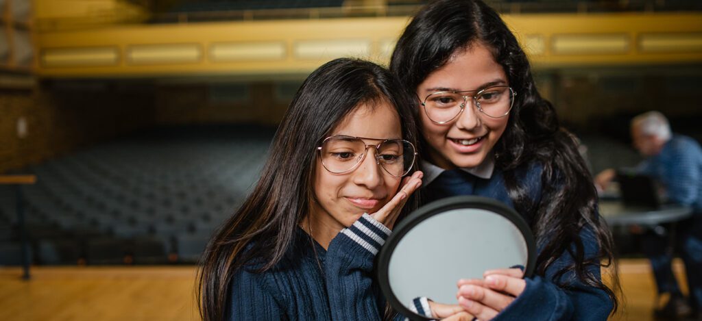 Two young girls wearing glasses closely looking at a round mirror in a large auditorium setting.