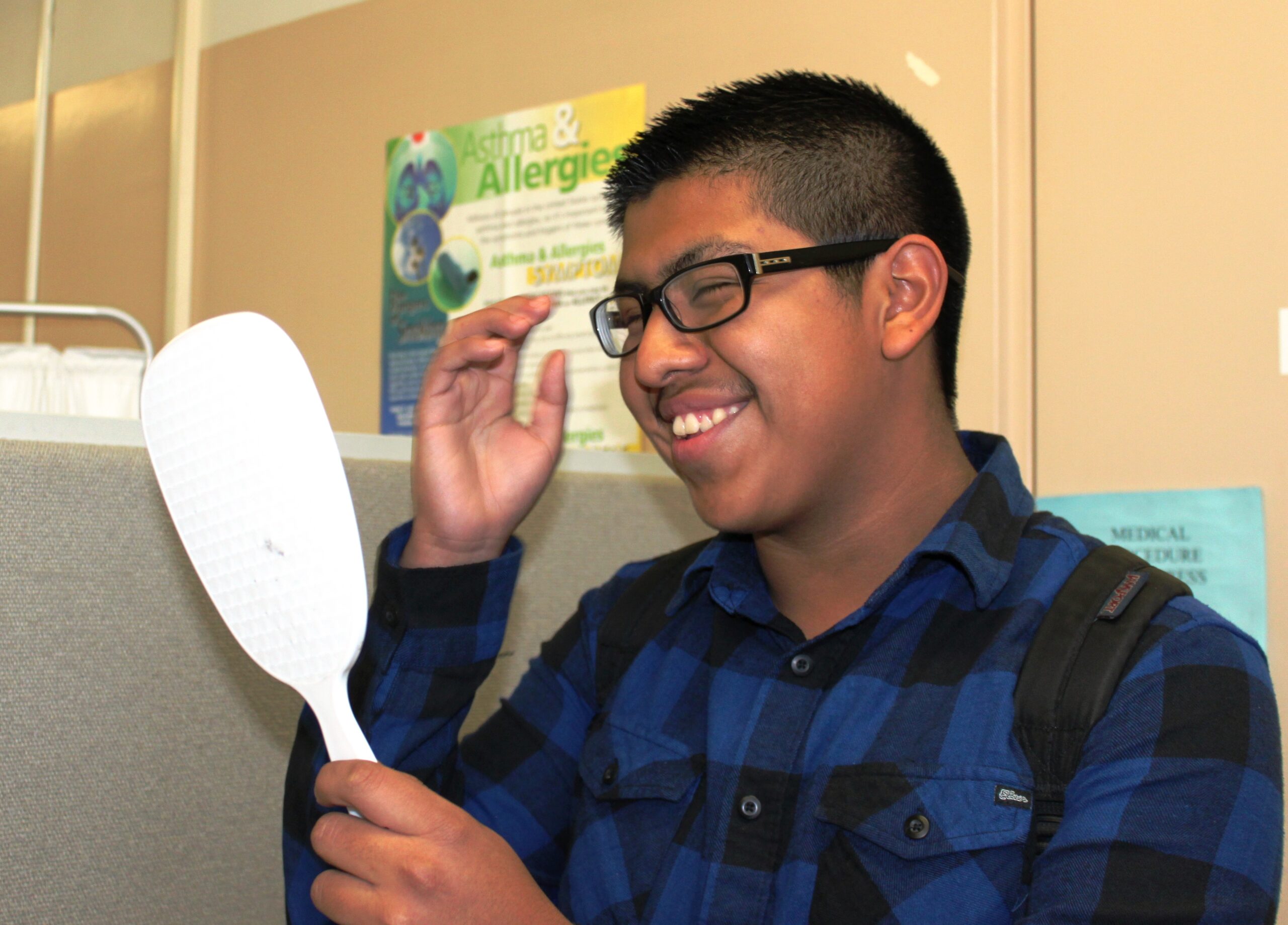 Smiling teenage boy wearing glasses and plaid shirt looks at himself in mirror.