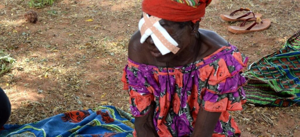 Woman with bandage over her eyes sits on ground outside in rural setting.