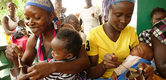 Two women sit closely feeding their infants with spoons, surrounded by other community members.