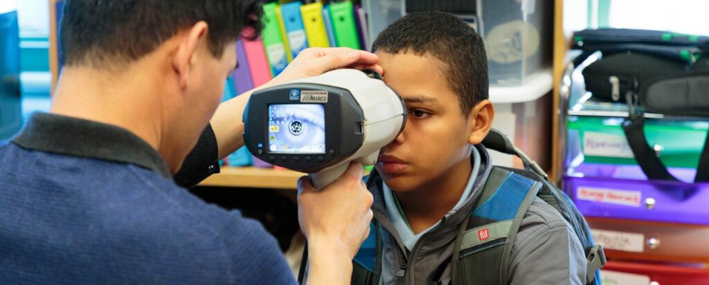 Young boy having his eyesight tested using a handheld vision screening device by a healthcare professional in a school setting.