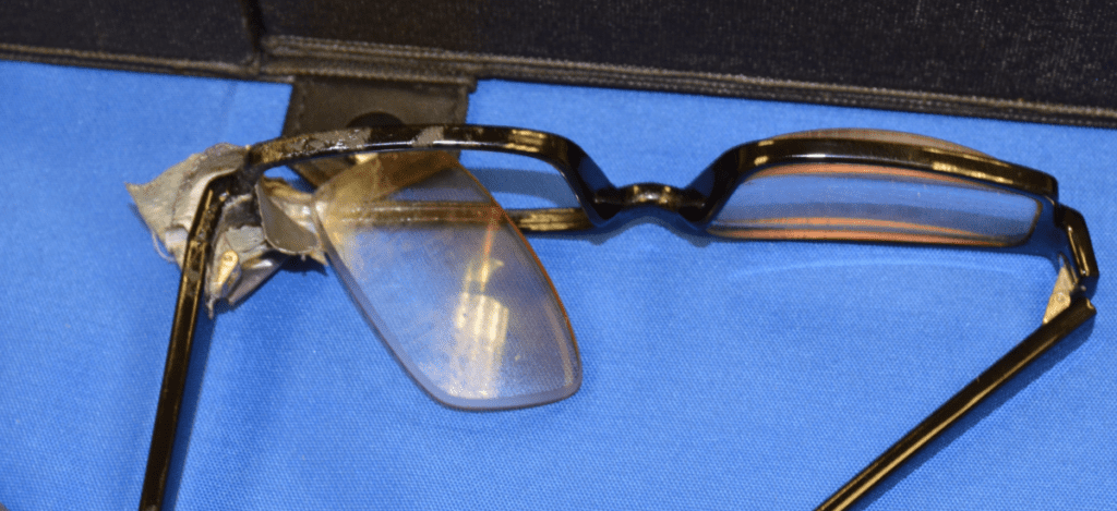 Close-up of damaged eyeglasses with a shattered lens resting on a blue cloth.