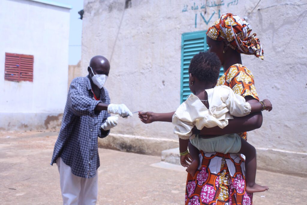 Masked healthcare worker hands vitamin A dose to a mother holding her a child in outdoor setting.