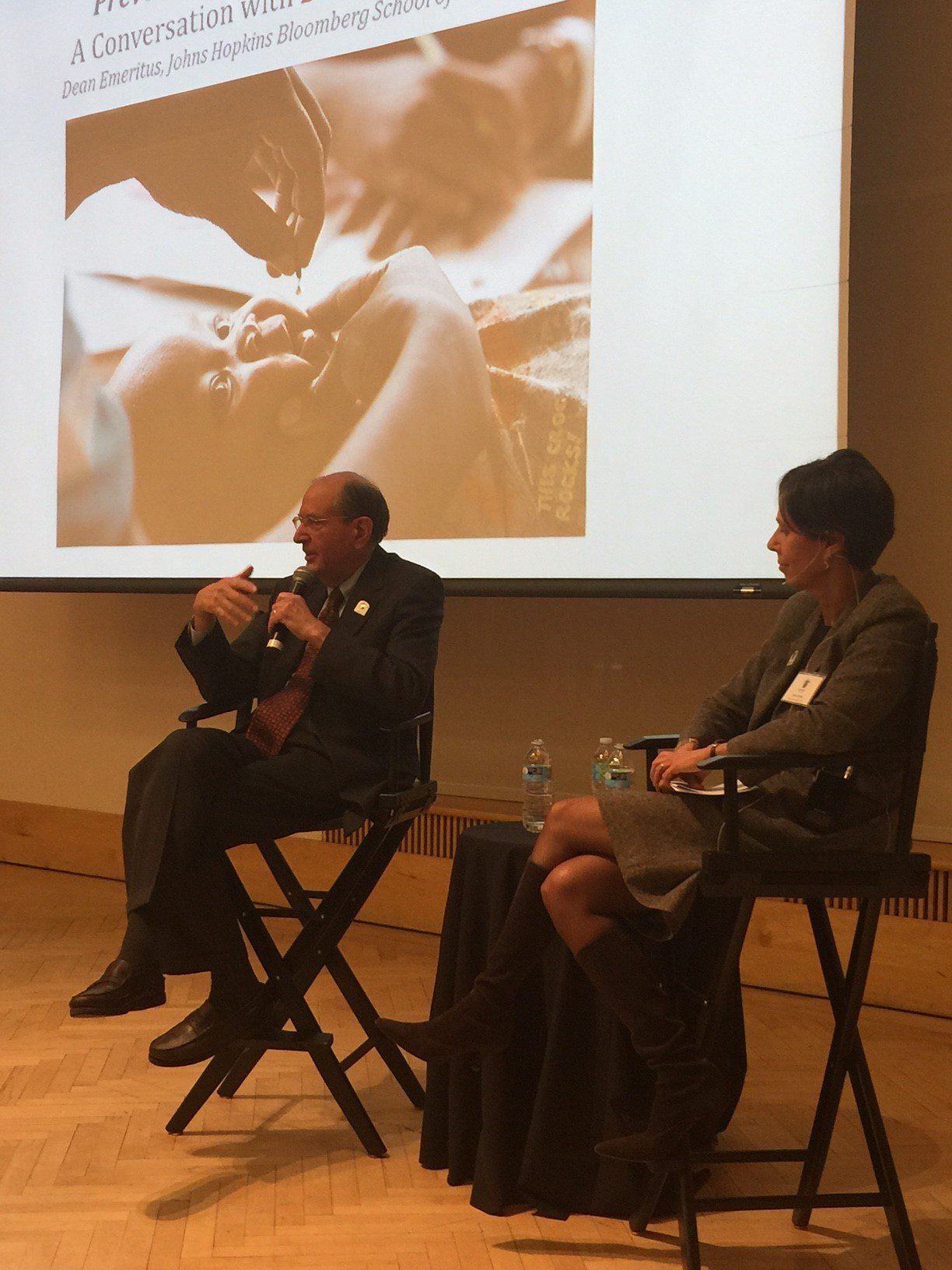 Man and woman seated on director chairs on stage, engaging in a conversation with a projected image of vitamin A supplementation behind them, in a formal event or conference setting