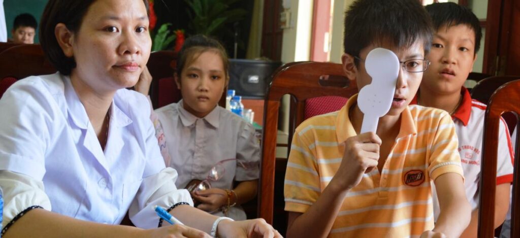 Young boy covers his eye during vision test with medical staff and other children in classroom setting.