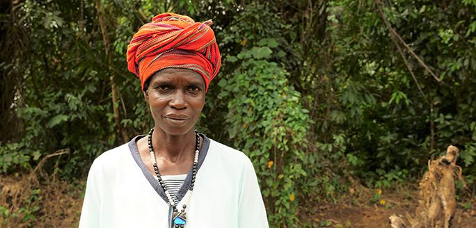 Woman with orange headwrap and white shirt standing in a green forest area.