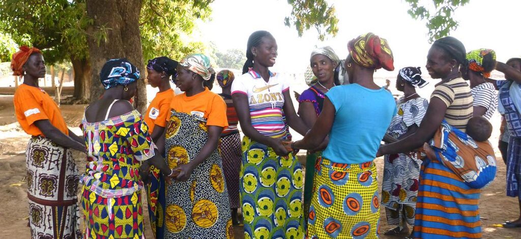 Several women wearing vibrant patterned skirts and headscarves gather under a tree, engaging in conversation in a rural setting.