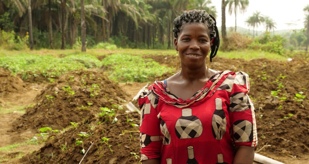 Smiling woman wearing a red patterned dress stands in a lush green farm field with palm trees in the background.