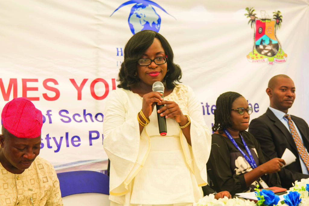Woman in white traditional attire holding microphone and addressing audience at an event.