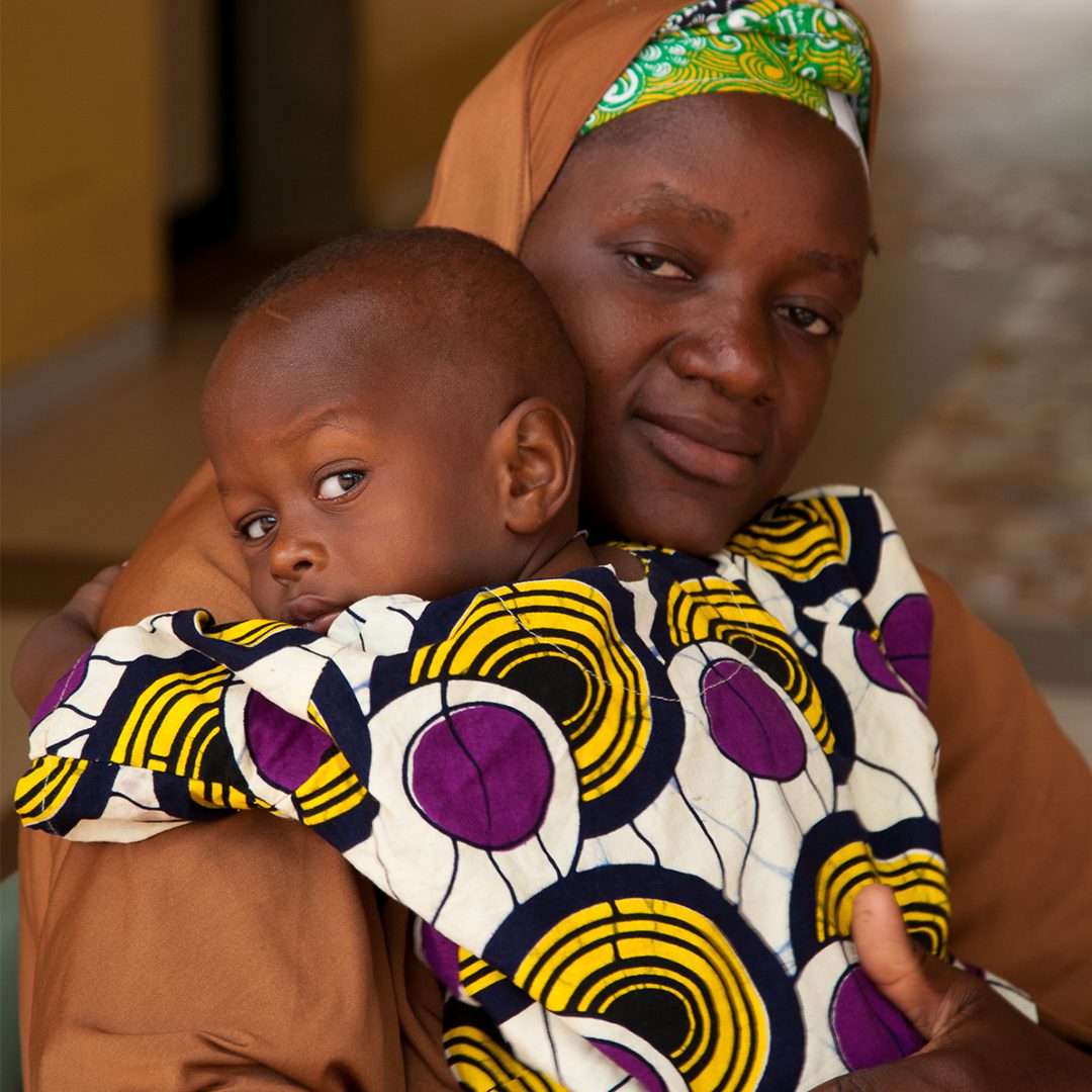 An African mother holding her infant son dressed in traditional clothing as they both look at the camera.