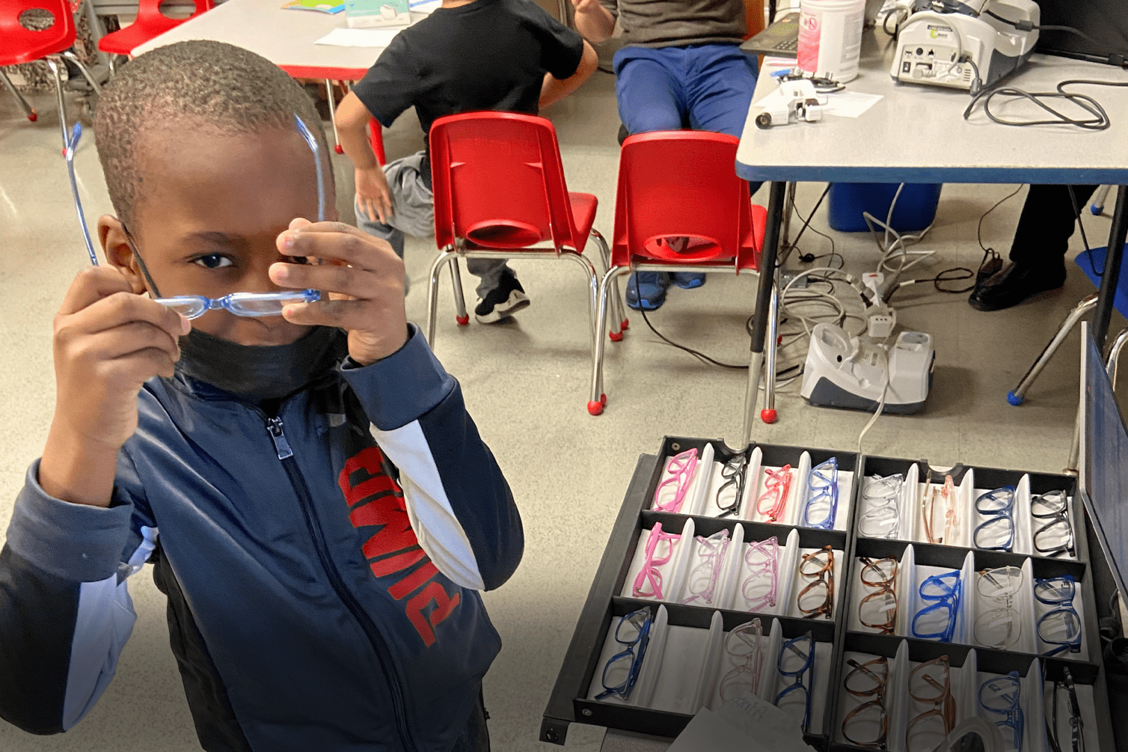 Prince, a young Black boy is in the middle of trying on a pair of blue rimmed glasses. He has his arms up and glasses more to the side as he's about to put them on. He's standing in front of a box with glasses in a classroom. He is wearing a black mask that is below his chin and a navy blue Puma zip-up hoodie.