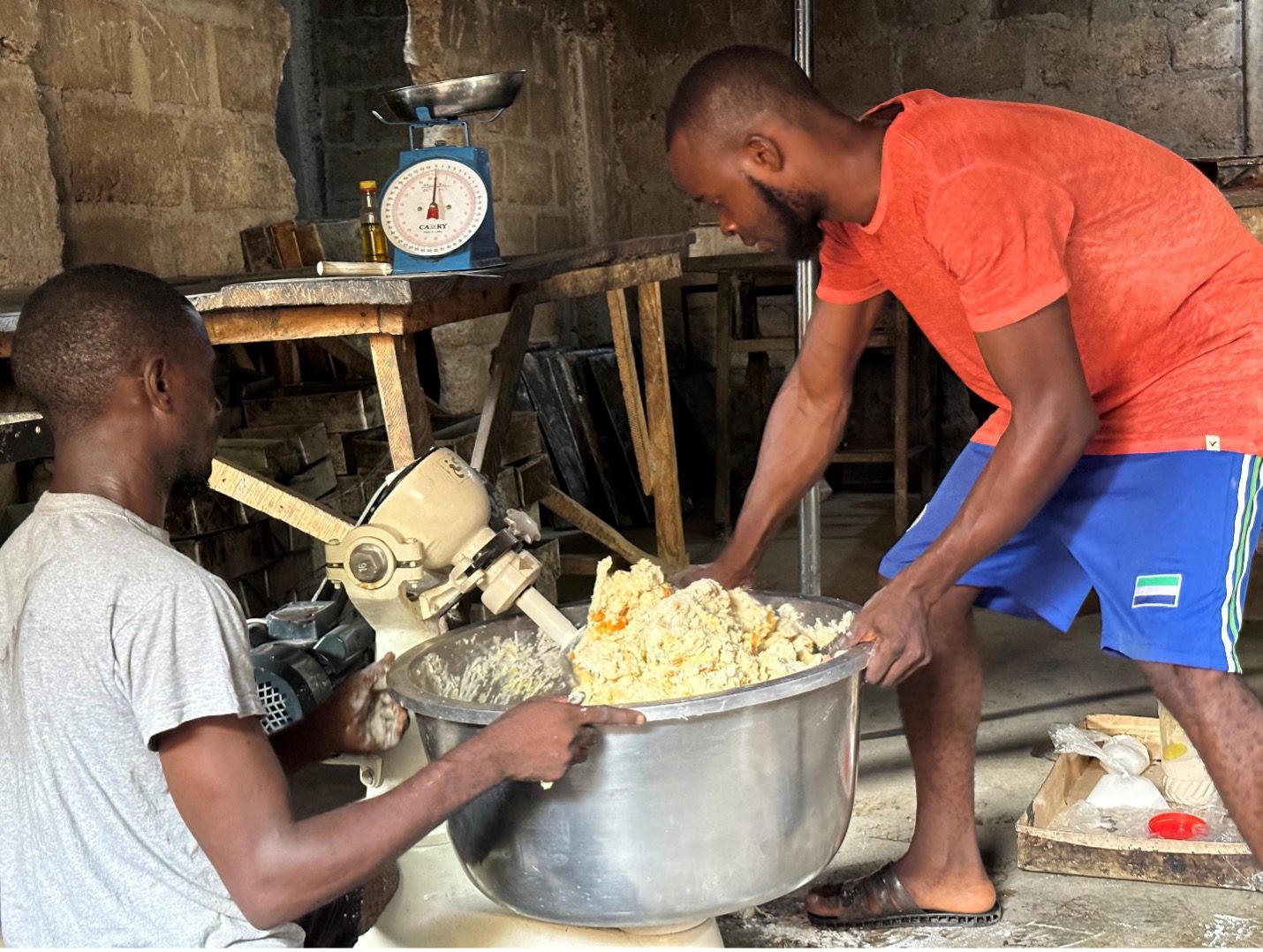 Two men with dark skin wearing t-shirts hold onto a large mixing bowl. Sweet potato bread dough is being mixed in the bowl with a large stand mixer. Behind them are work benches.