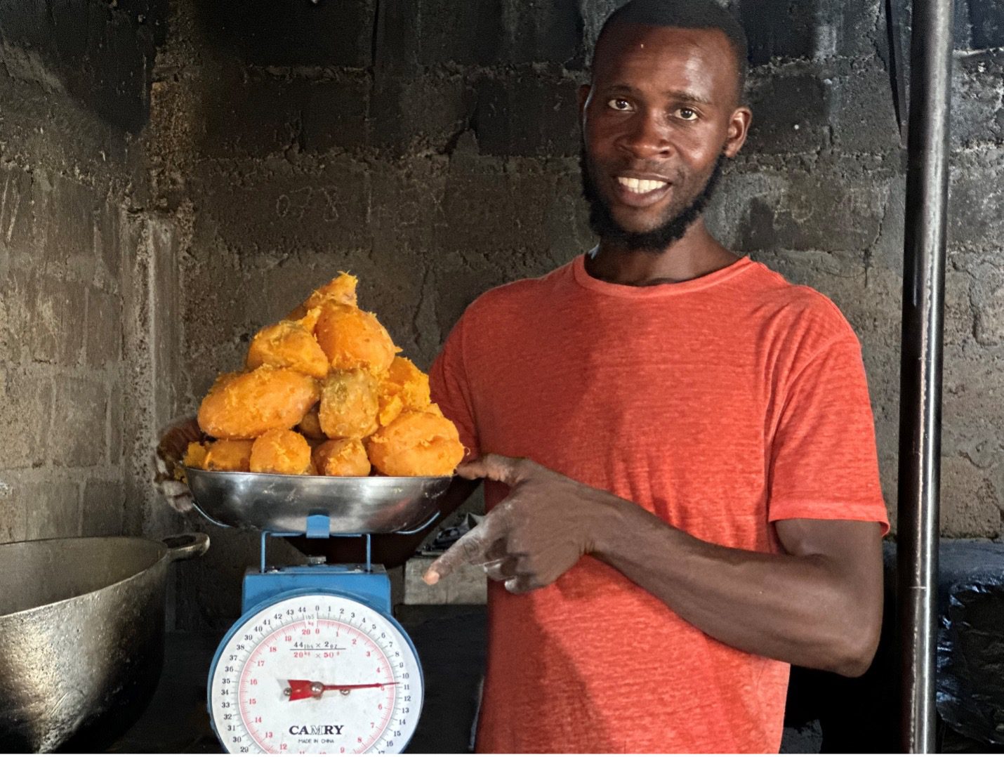 A man with dark skin wearing a red t-shirt holds a scale with a pile of cooked sweet potatoes on it.