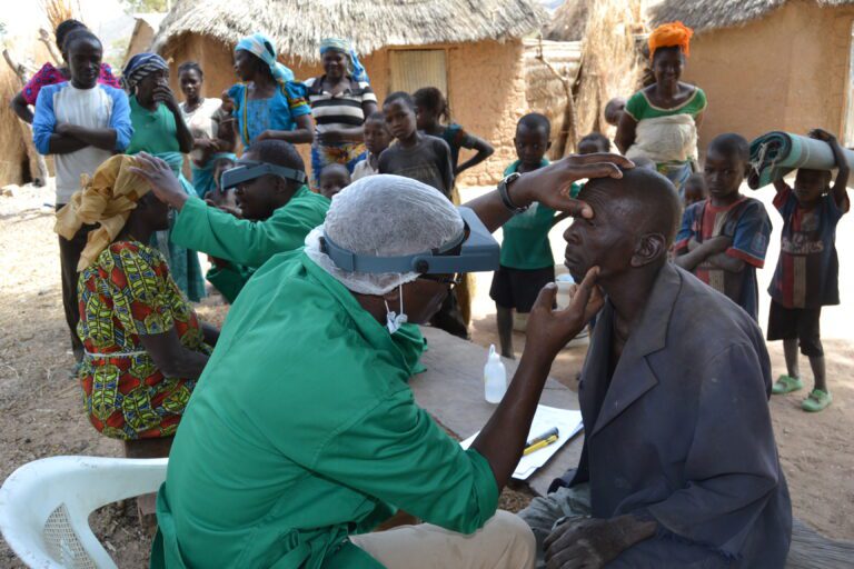 Medical professional in protective gear checking a man's eye outdoors in a rural community, with people in background.
