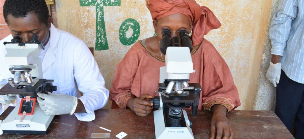 Woman in traditional attire and man examining samples under microscopes in an outdoor rural clinic setting.