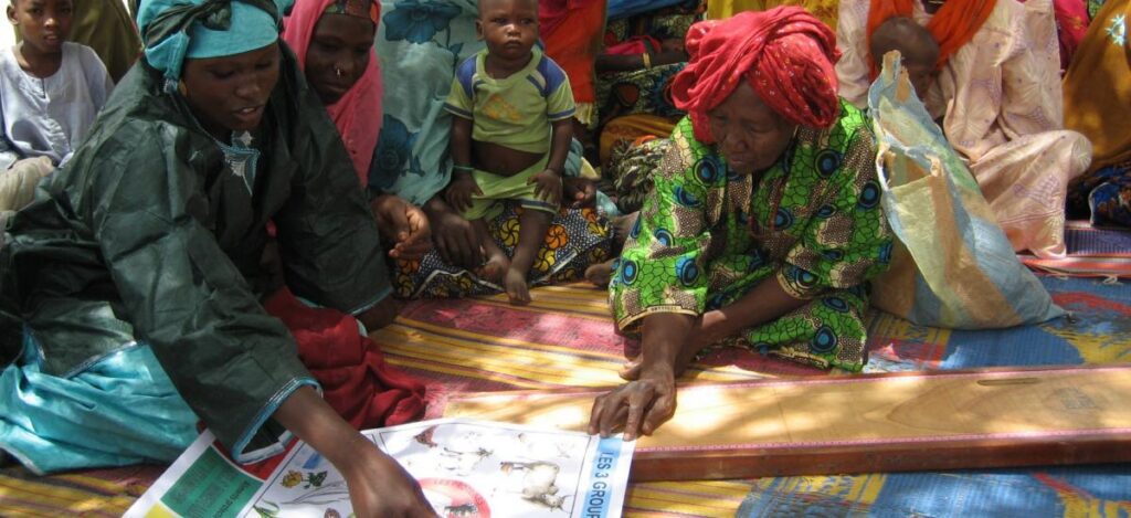 Group of women and children gathered on mats in outdoor rural setting, interacting with printed learning resources.