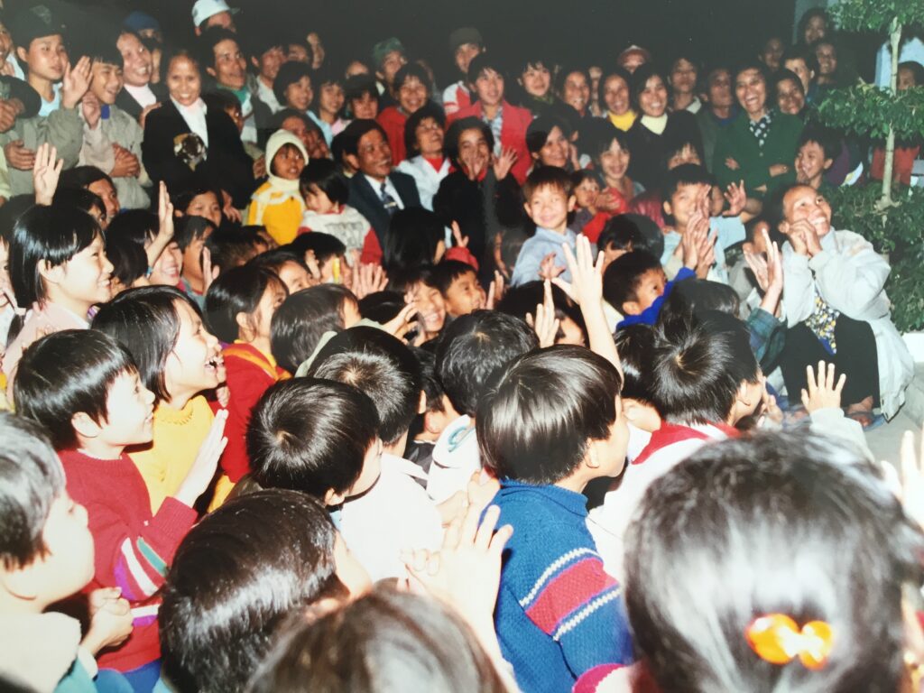 Numerous children gathered closely, sitting and clapping during an indoor event.