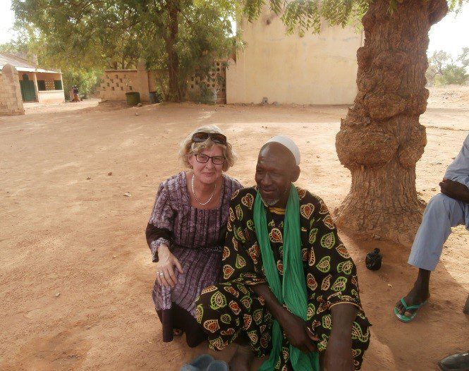 Smiling woman kneeling next to elderly man wearing patterned clothing and green scarf under a tree in a rural village setting.
