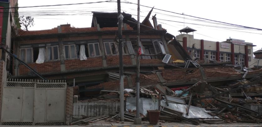 Severely damaged multi-story building collapsed with rubble and broken wooden beams, showing the aftermath of a natural disaster.