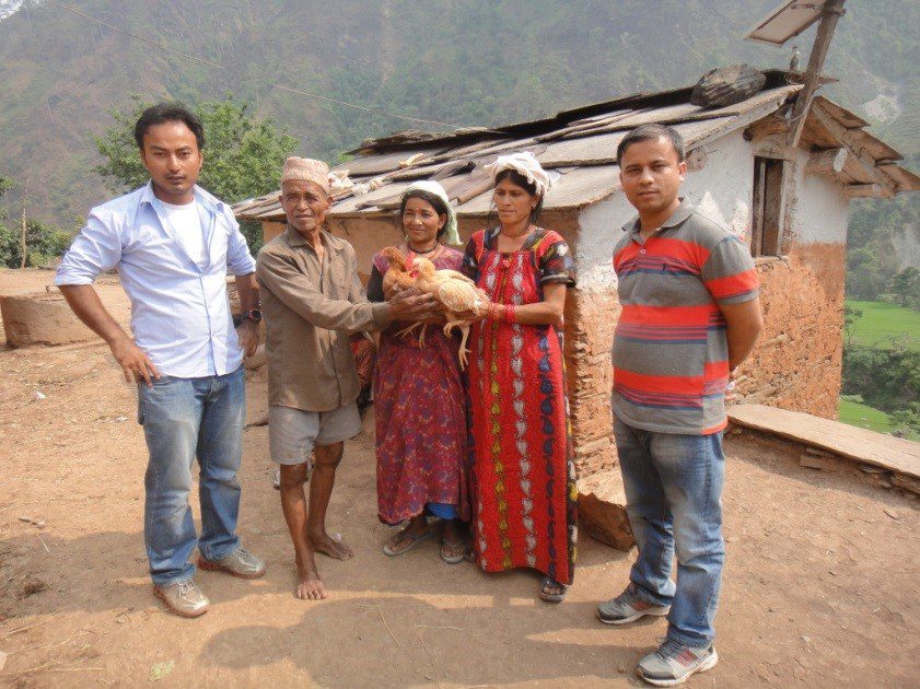 Five rural individuals stand on a dirt path in front of a rustic house, with one woman holding a chicken.