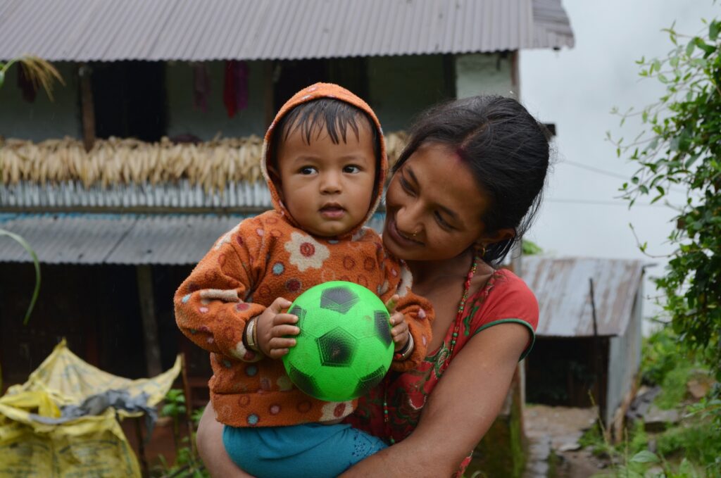 Smiling mother holds toddler wearing orange hoodie and blue pants, who is holding a green and black soccer ball. The scene is set outside a rustic home with greenery around.