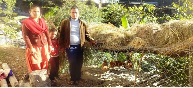 Man and woman standing next to a large bundle of dried hay in a garden area with plants and greenery, showing agricultural activity and rural lifestyle outdoors