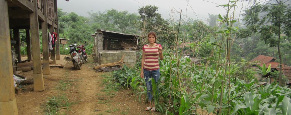 Girl in striped shirt standing on dirt path beside garden with green plants and rustic wooden buildings in background.