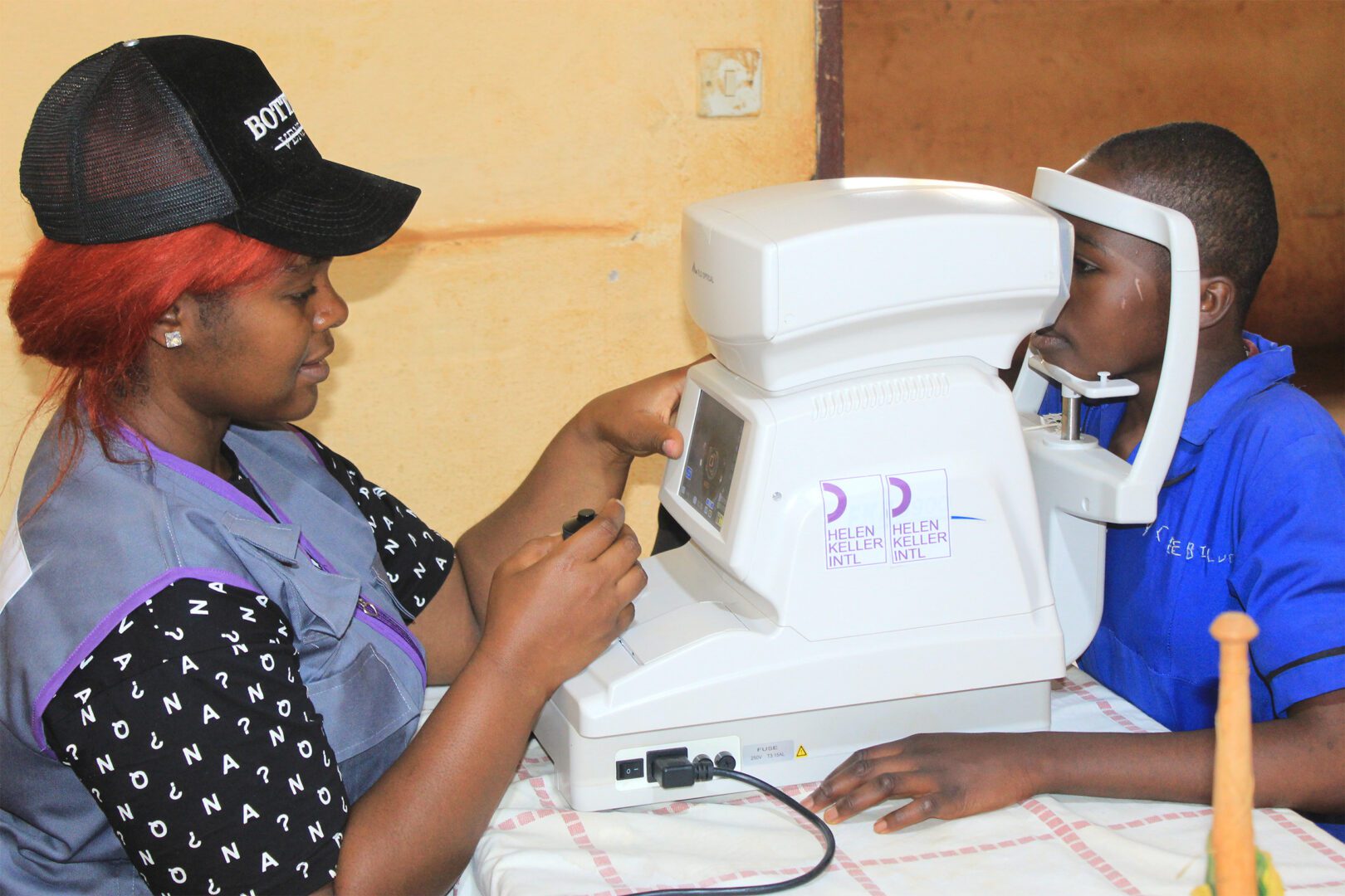 A young girl with dark skin wearing a blue top sits with her face in an autorefractor. A woman with dark skin and bright red hair wearing a black cap, grey and purple vest, and black shirt with white letters on it uses the machine to examine her eyes.