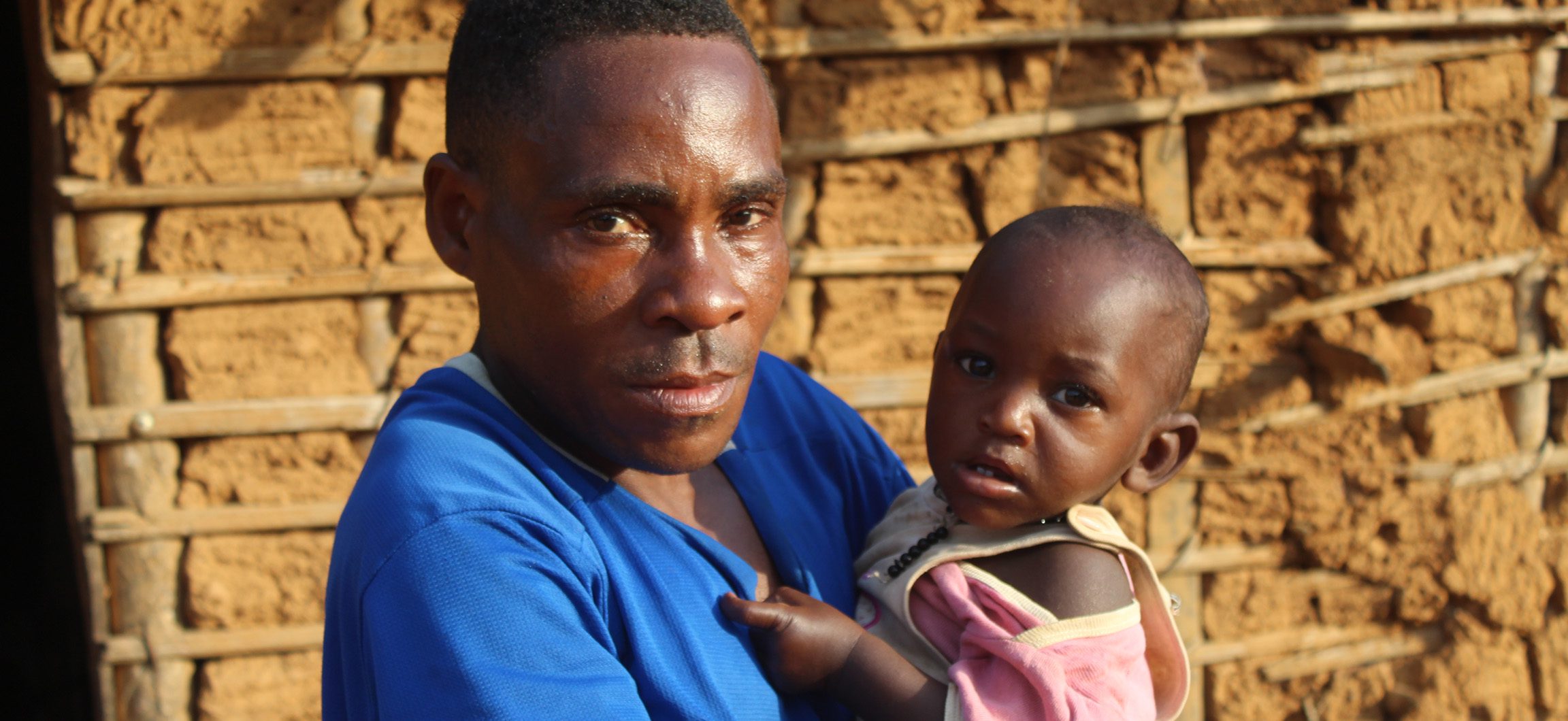Father in Cameroon holds his young baby in his arms while standing in front of his home.