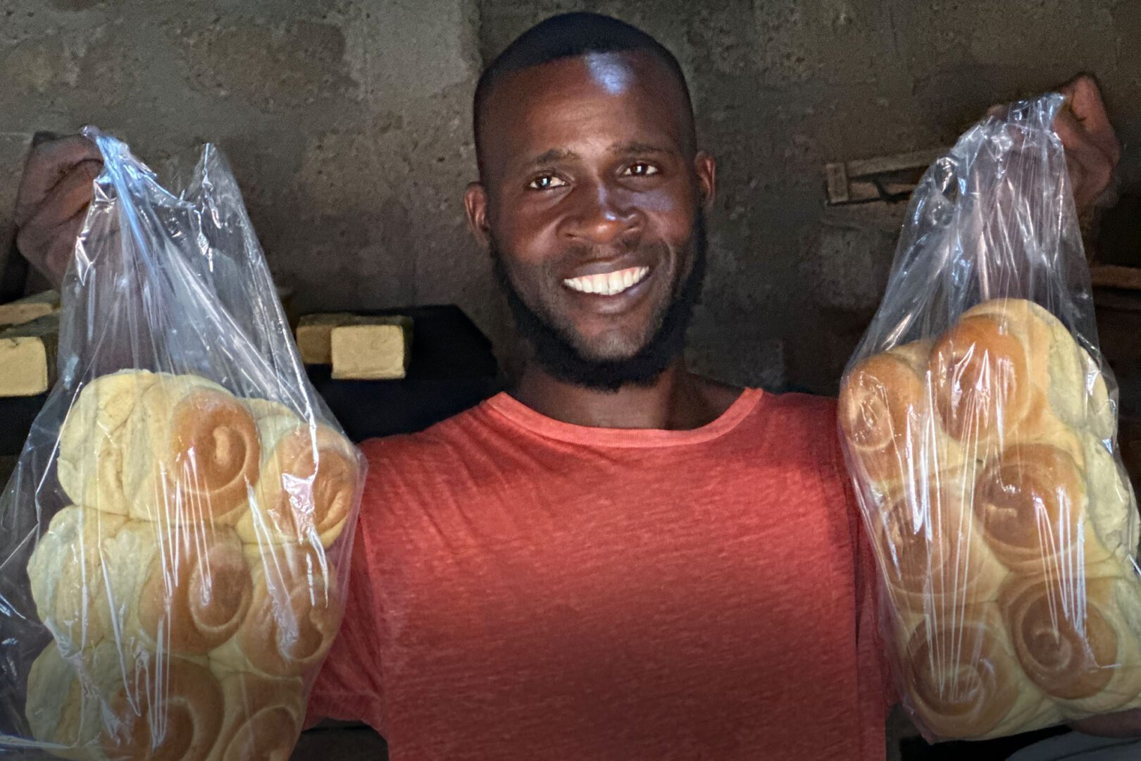 African man from Sierra Leone holds up baked breads he made while smiling.
