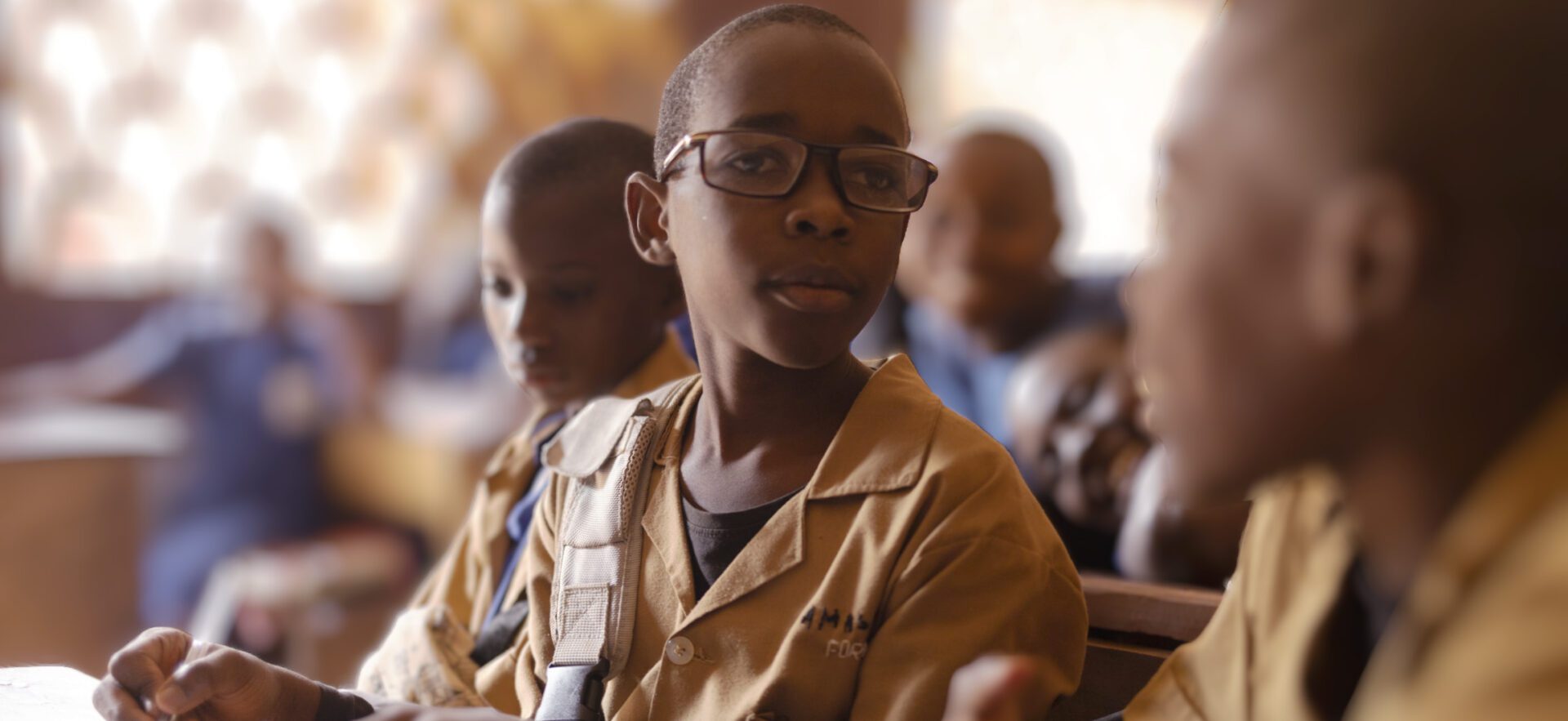 A boy with dark skin and glasses sits at a desk in a classroom surrounded by peers. He is turned toward the camera.