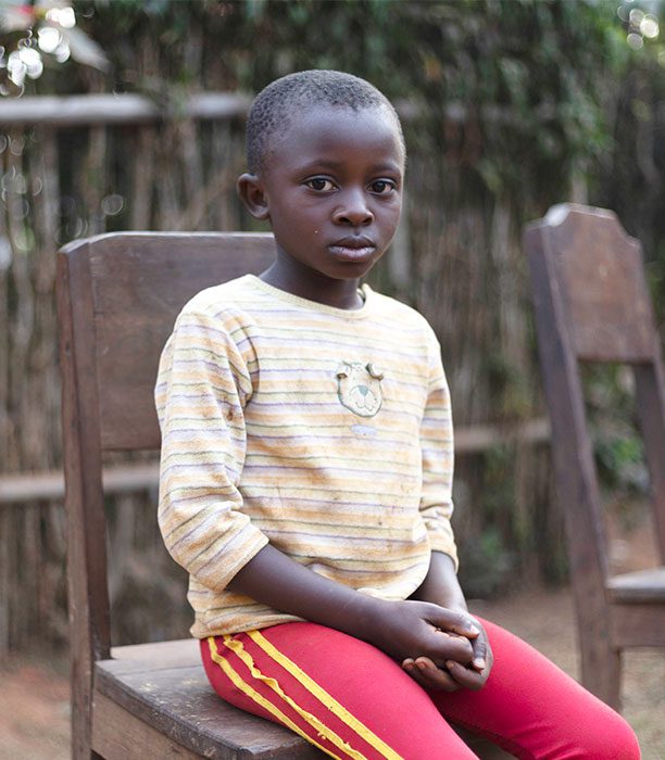 Boy in Cameroon sitting on a wooden chair wearing a yellow shirt and red pants looks at the camera with a neutral face.