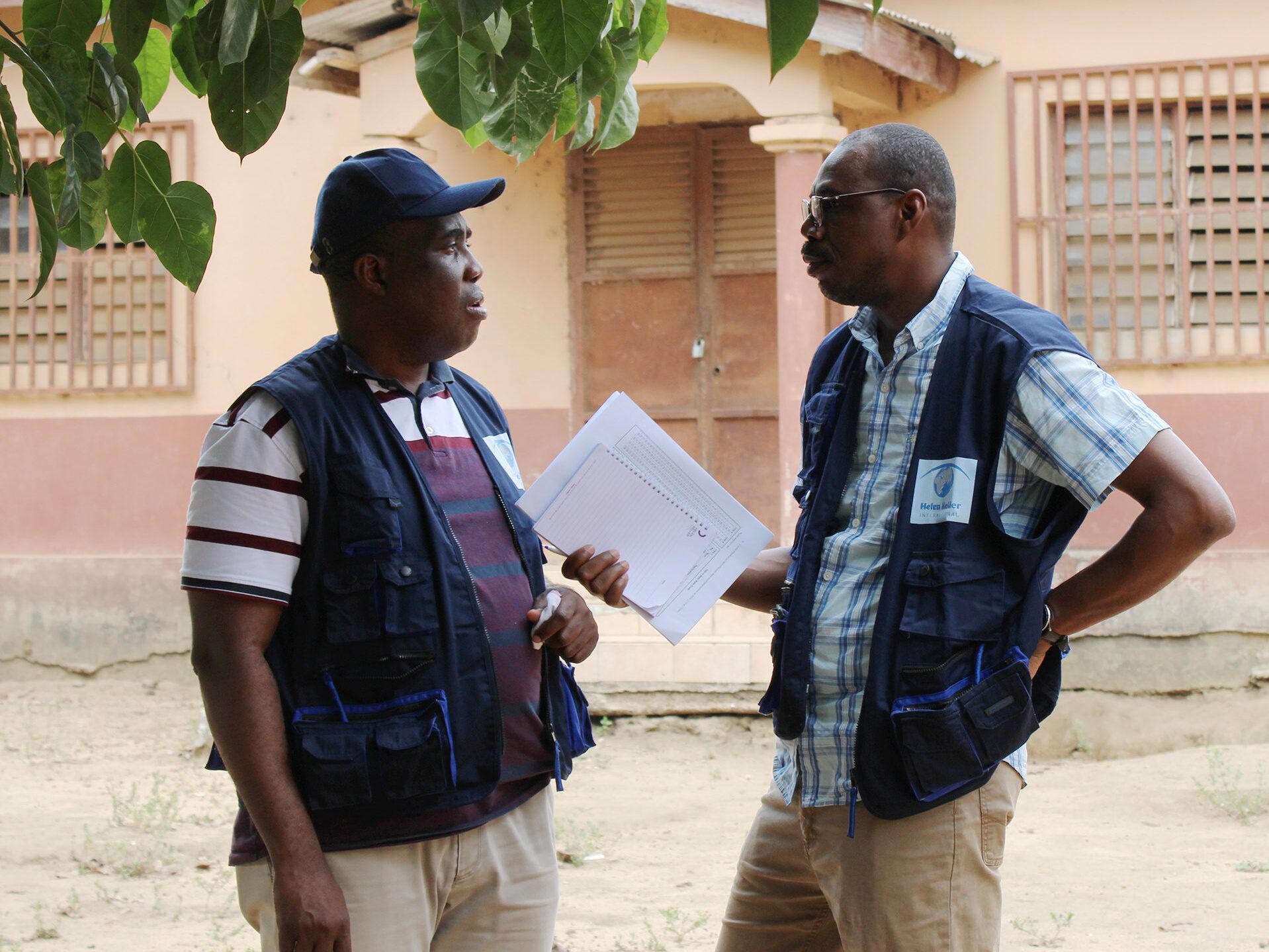 Two men with dark skin stand next to each other speaking outside. The man on the right is holding a notebook and papers and gesturing as he speaks.