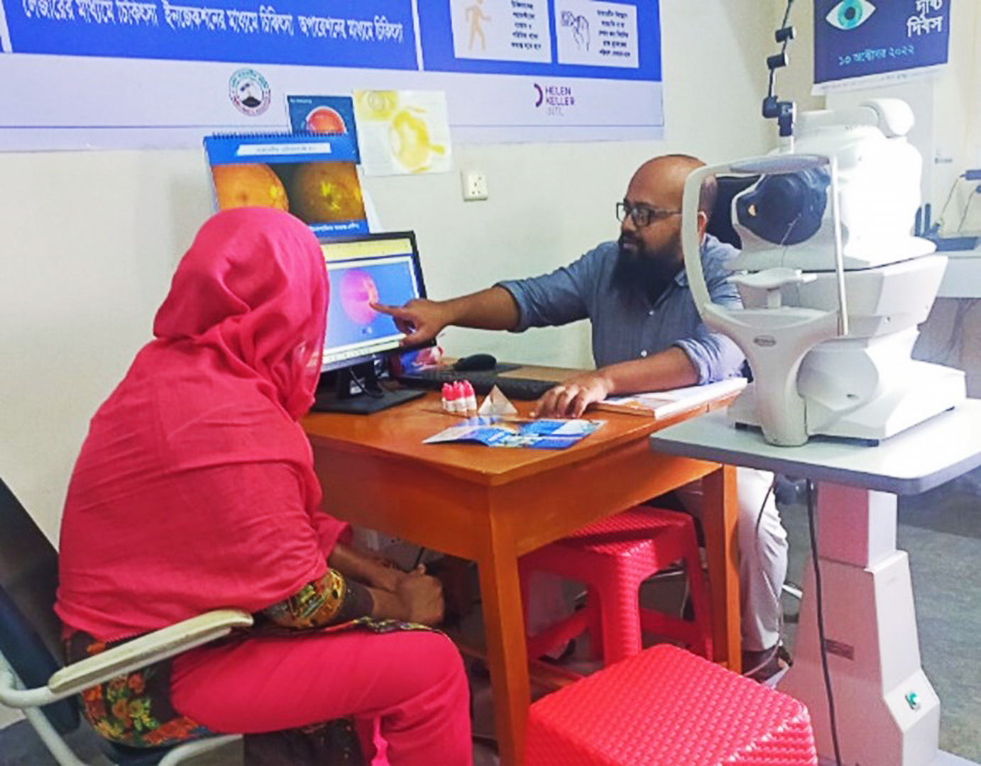 A woman in a bright pink sari sits across the table from a bald man wearing glasses. The man points to an image of a retina on a computer screen.