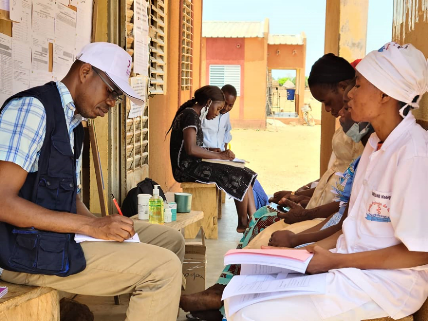 A man sits across from a female health worker outside of a building. The man is writing in a notebook.