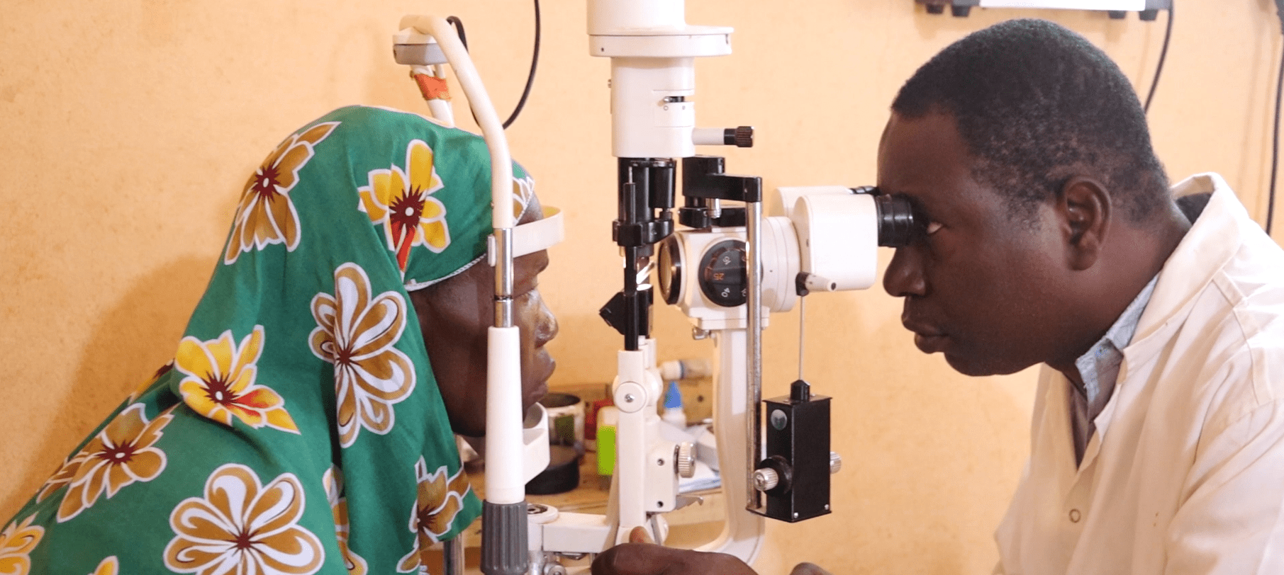 A woman wearing a headscarf with a flower pattern has her eyes examined by a healthcare worker.