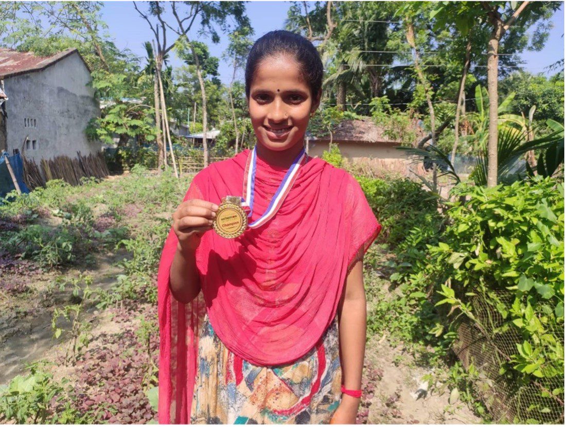 A teenager from Bangladesh shows off a gold medal hanging around her neck. She is standing outside with greenery behind her.